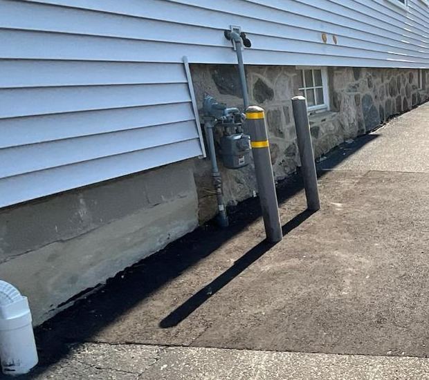 An exterior gas meter mounted on a house wall with two concrete filled bollards protecting it from the driveway